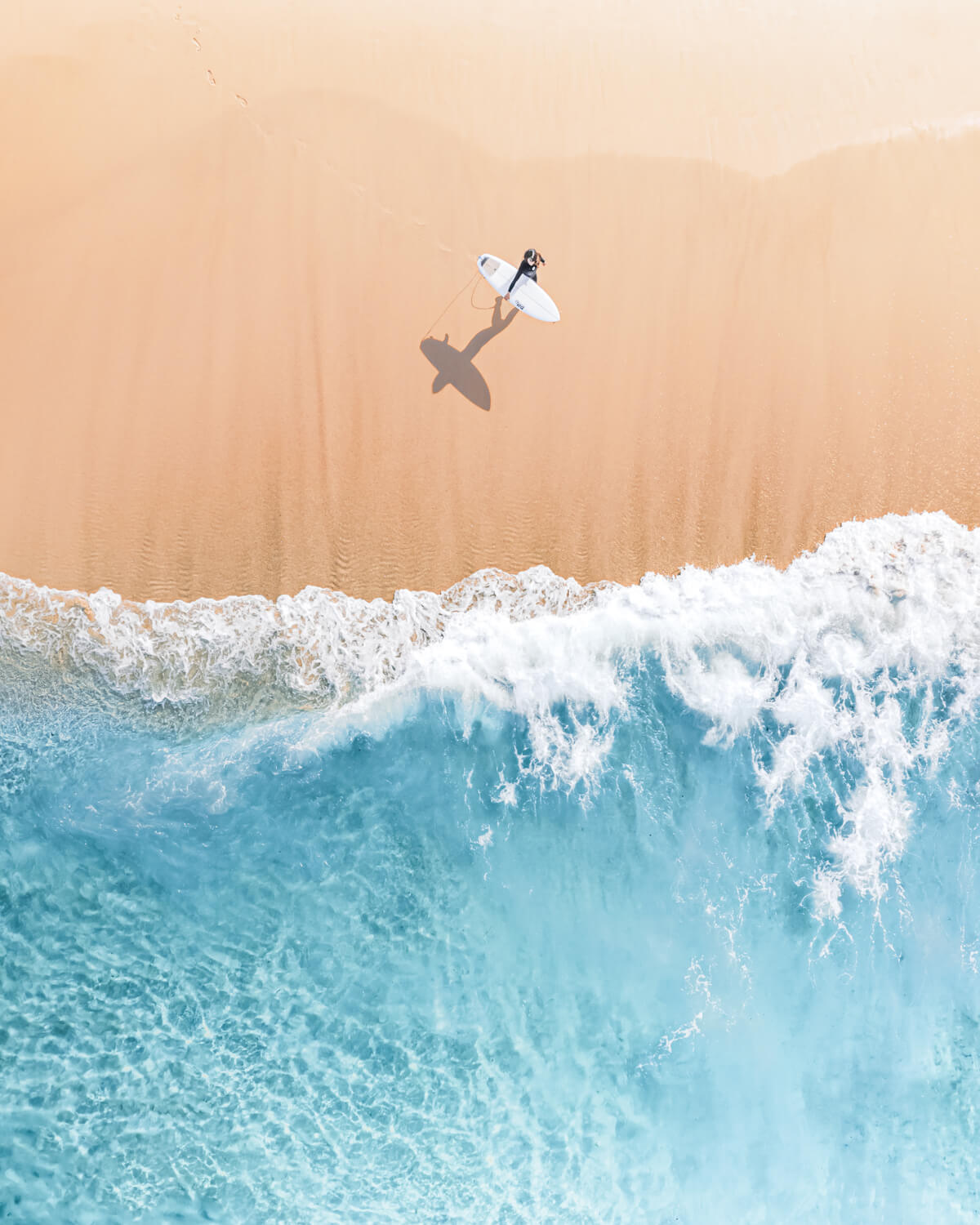 Aerial view of a lone surfer walking toward the breaking waves at Bronte Beach, Sydney