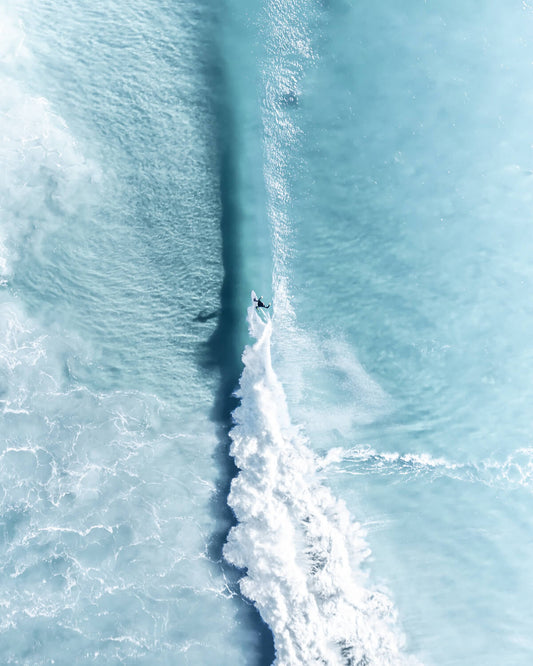 Aerial view of a lone surfer leaving a vertical white wake through calm turquoise water at Byron Bay