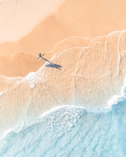 Aerial view of a lone surfer carrying a board and casting a long shadow on golden sand at The Pass, Byron Bay