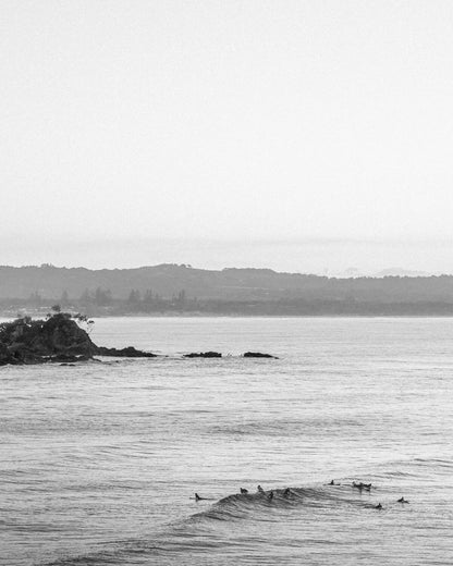 Black and white aerial view of surfers riding a wave at Wategos Beach, Byron Bay with the headland behind