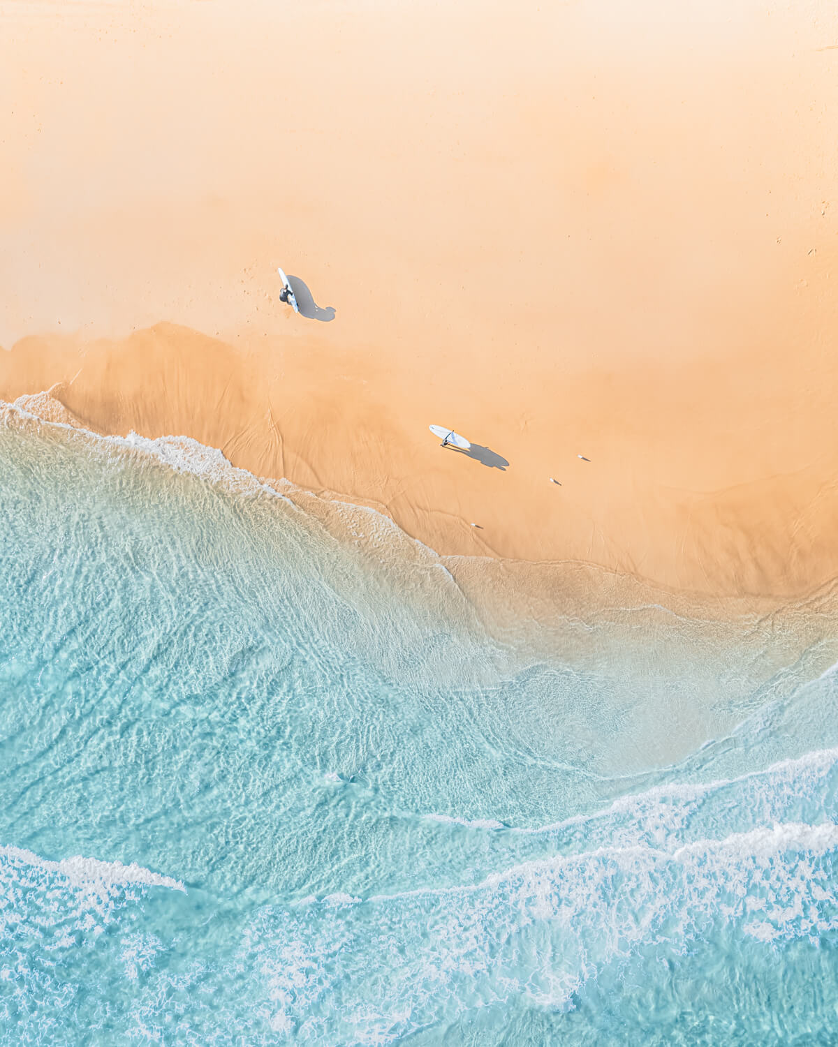 Aerial view of two surfers carrying boards and casting long shadows on golden sand at The Pass, Byron Bay