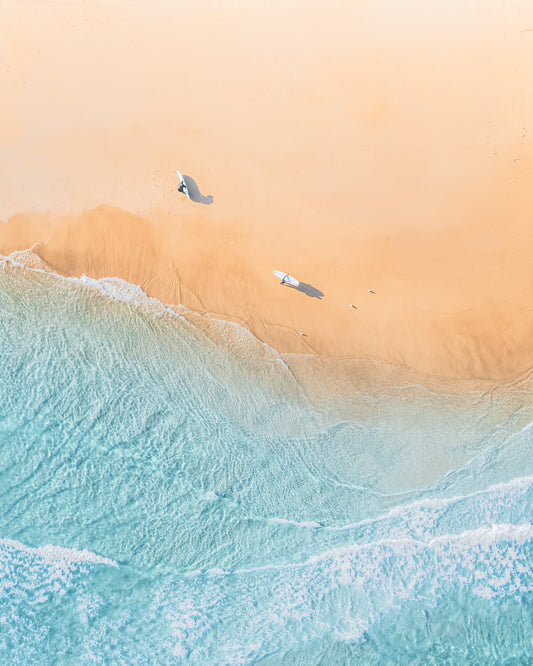 Aerial view of two surfers carrying boards and casting long shadows on golden sand at The Pass, Byron Bay